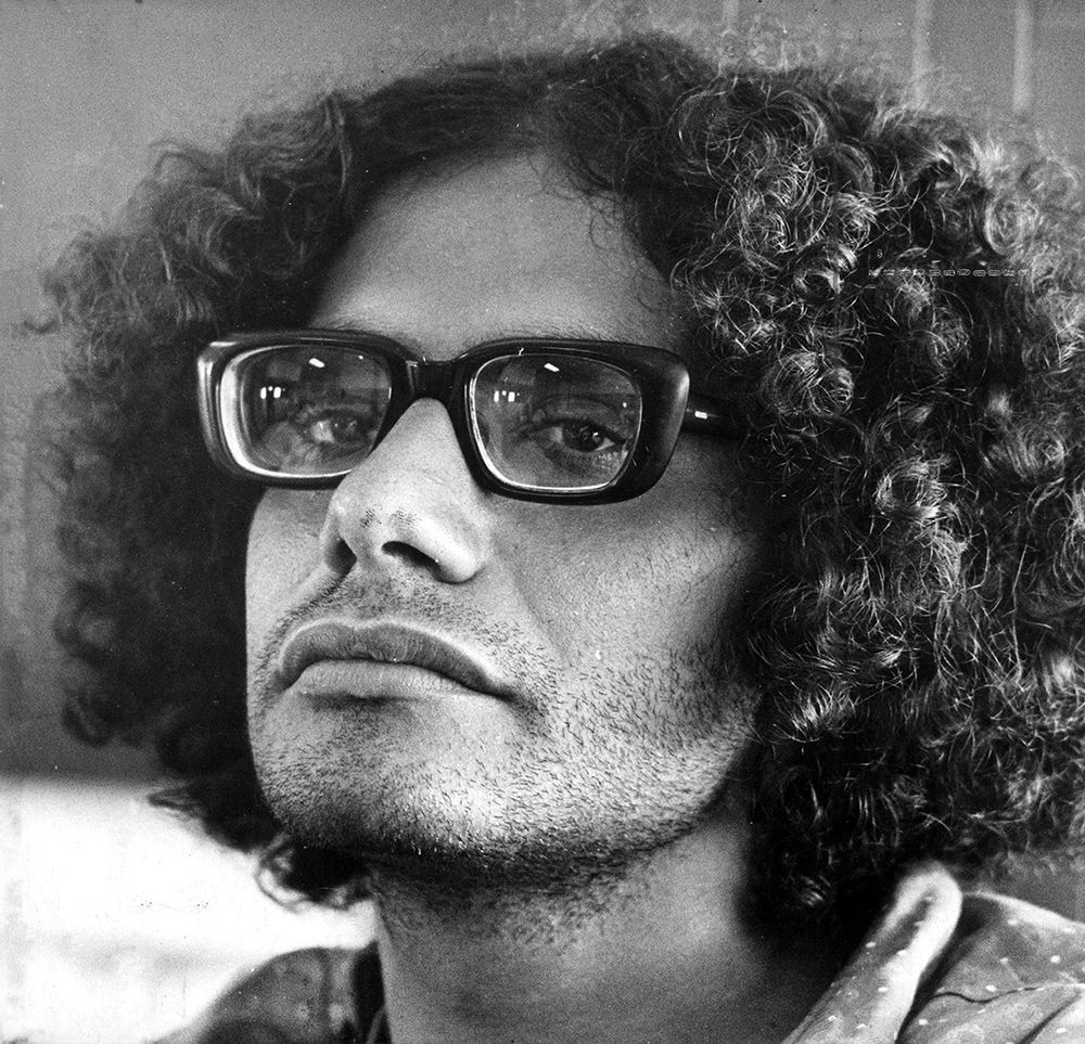 Imagem de perfil do poeta. Ele tem cabelo crespo volumoso, usa um óculos com aros pretos grossos e possui uma barba rala, com cavanhaque.