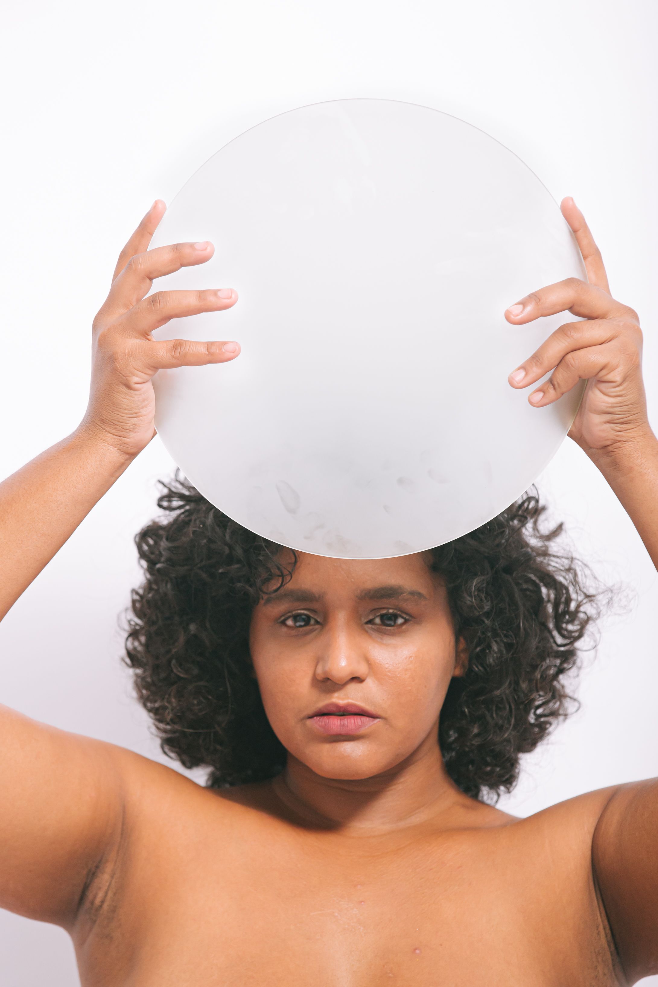 Mulher afro-indígena, com pele de cor amendoada, aparece de pé, com o dorso nu. Ela está séria e segura uma peça redonda e branca acima da cabeça. Seus cabelos são crespos e escuros, na altura dos ombros. 