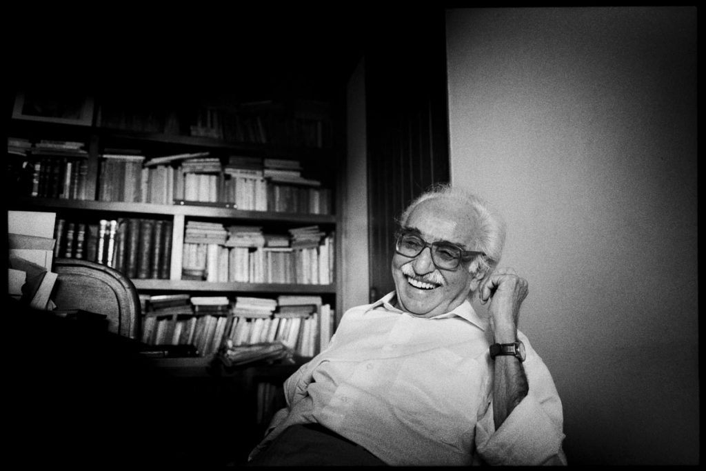 Manoel de Barros aparece sentado, sorrindo, vestindo uma camisa e usando óculos e relógio. Atrás dele, há uma estante cheia de livros.