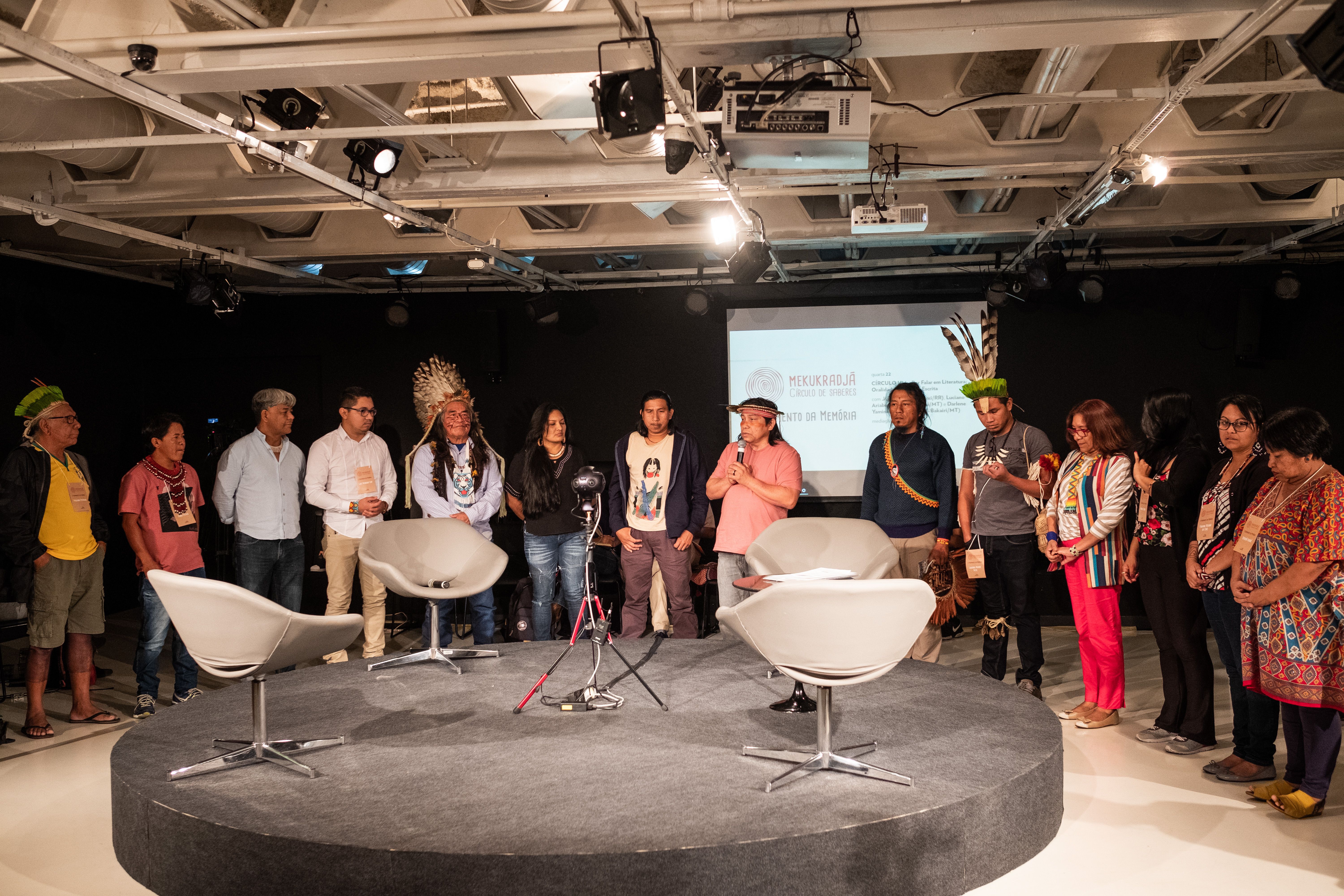 Palco redondo e baixo cinza, com quatro cadeiras brancas giratórias vazias em cima. Ao redor, 14 pessoas, entre homens e mulheres, todas indígenas, aparecem de pé. A maioria das mulheres está à direita da imagem e a maioria dos homens está à esquerda. Ao centro, um dos homens está com um microfone na mão.