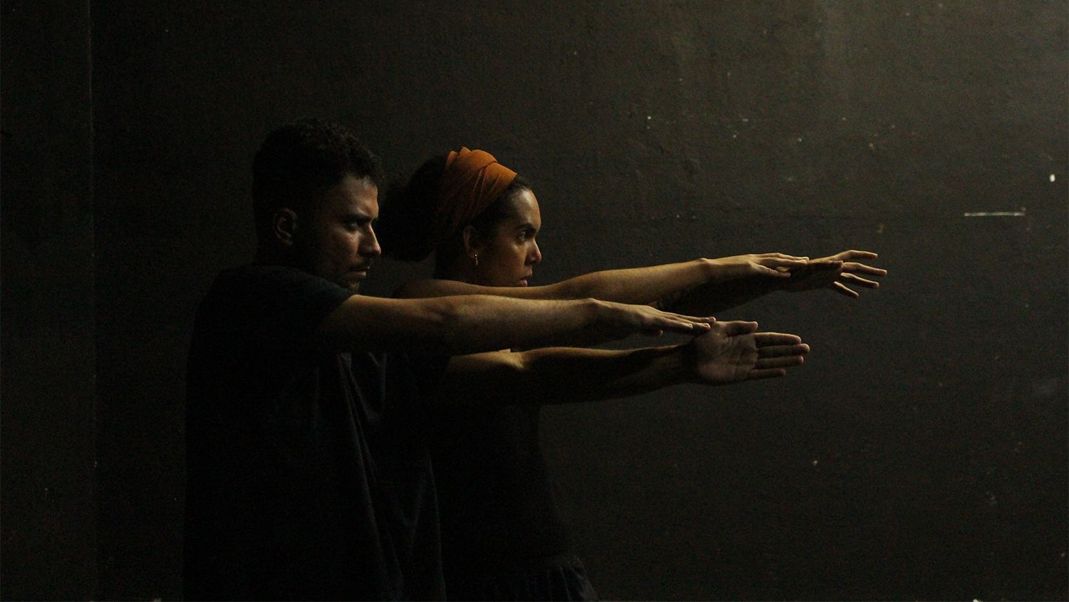 Fotografia colorida do espetáculo Elefante. Na cena, um ator e uma atriz estão de perfil com os braços esticados na horizontal, apontados para a direita.