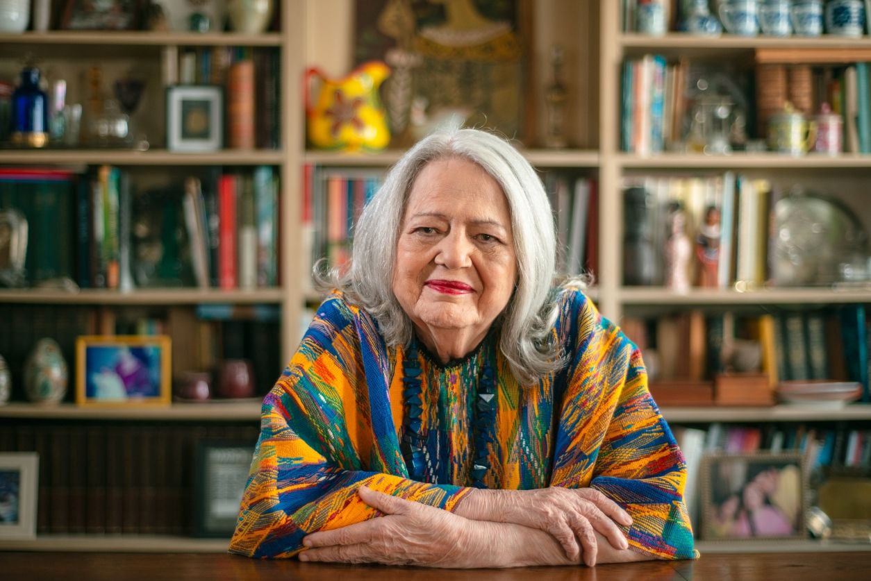 Fotografia colorida de Ana Mae Barbosa. Ela é uma mulher branca, idosa, possui cabelos brancos. Está usando uma roupa colorida, com tons de laranja, azul e verde, e um colar azul. Fotografia colorida de Ana Mae Barbosa. Ela é uma mulher branca, idosa, possui cabelos brancos. Está usando uma roupa colorida, com tons de laranja, azul e verde, e um colar azul.