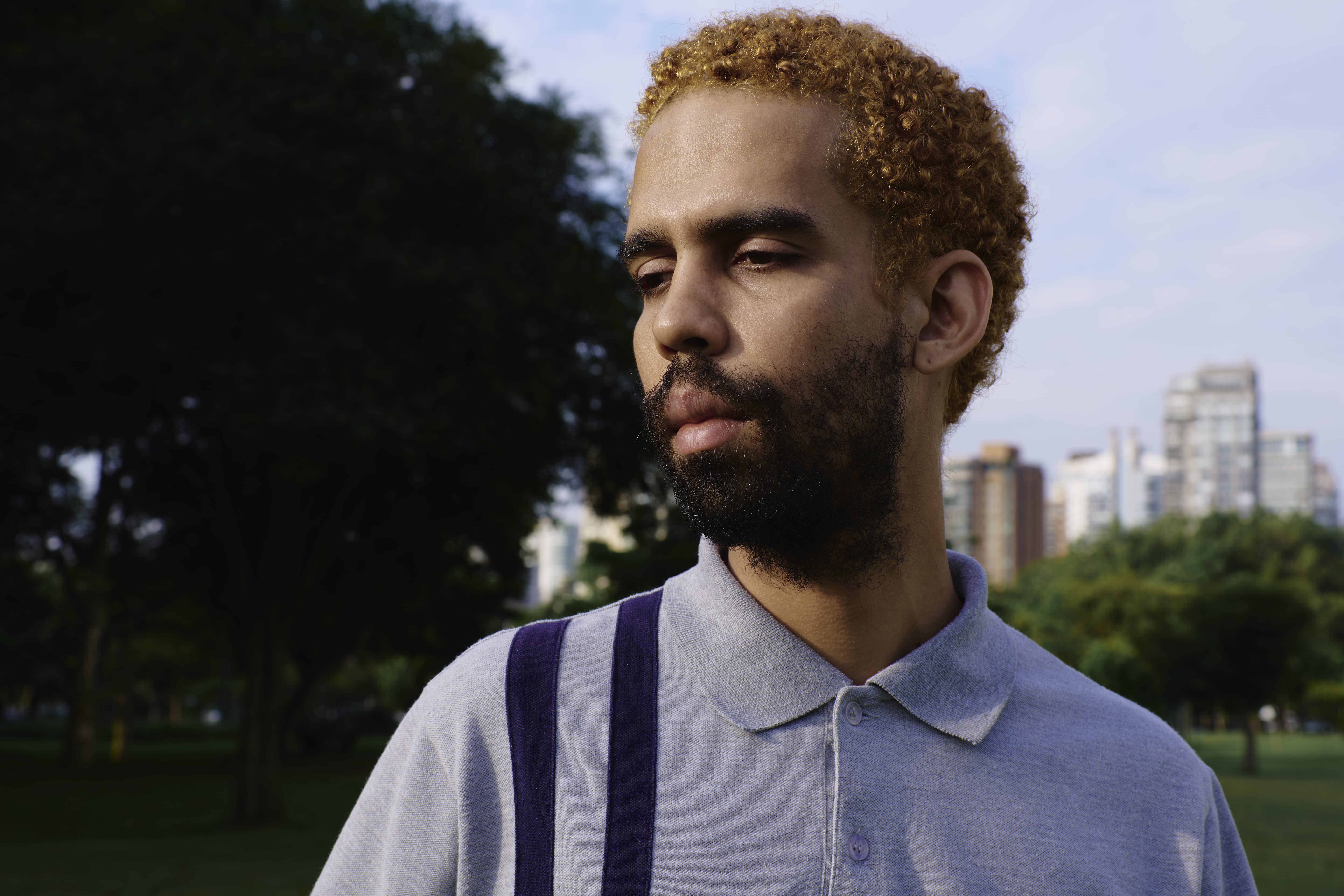 Homem jovem e negro com a pele clara aparece em foto dos ombros para cima, em parque. Ele usa camiseta pólo cinza, com duas listras azuis verticais. Ele tem barba escura e os cabelos crespos e curtos pintados de louro escuro.