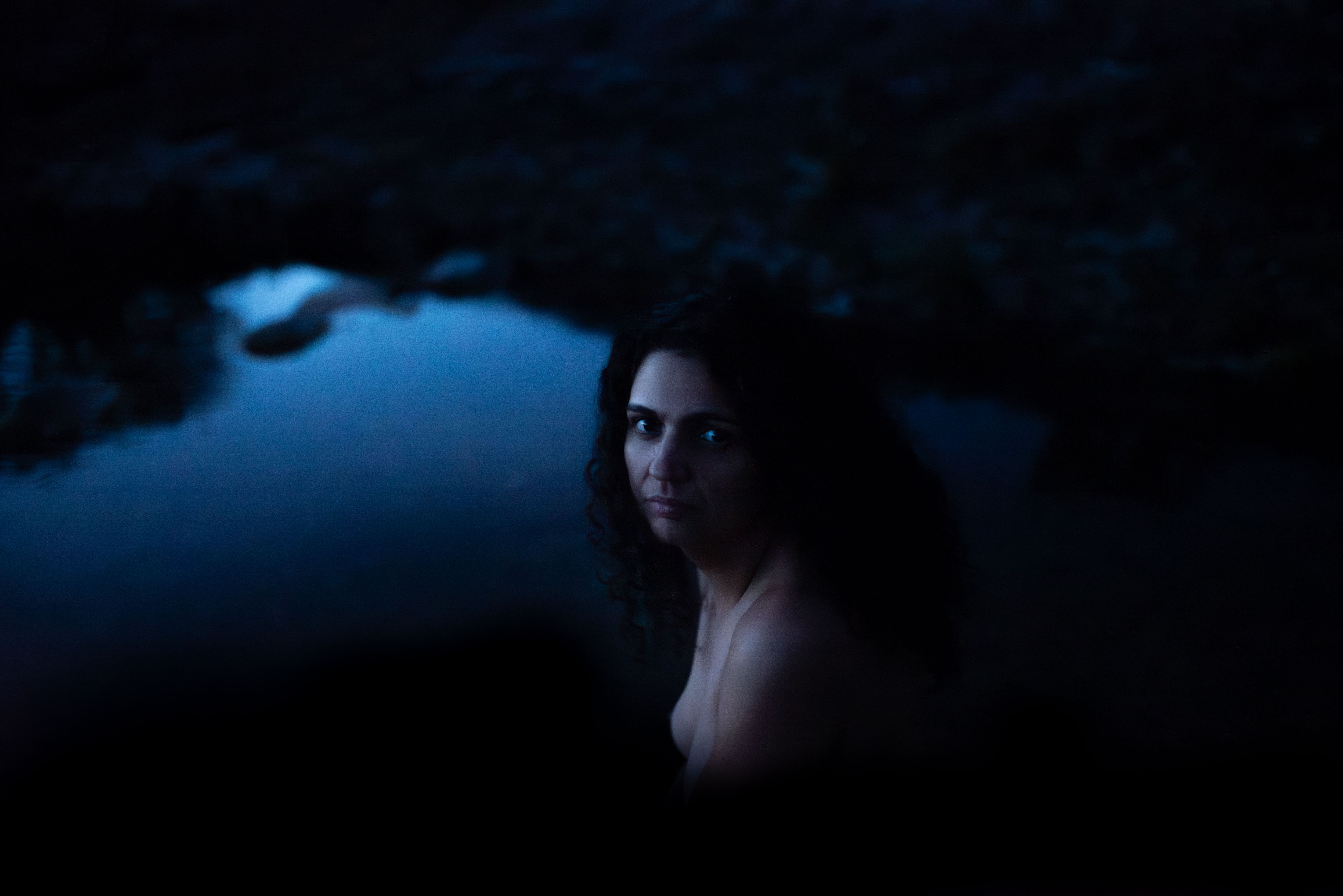 Luiza Brina aparece perto de águas. Ela está nua e olha séria para a câmera. A artista tem cabelo encaracolado. A imagem trabalha com sombras e falta de luz.