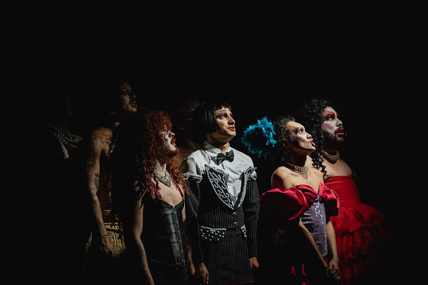 Fotografia colorida de cena do espetáculo Sebastião. Na imagem, sete pessoas vestida como drag estão com o corpo virado para a direita e olha para cima.