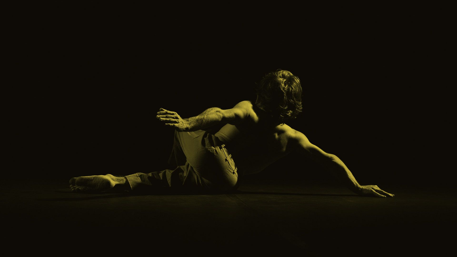 Fotografia do ator Giovanni Venturini em cena. Na imagem, que possui um filtro amarelo, característico do podcast Ficções, o ator performa seu espetáculo no chão de um palco, com os braços esticados. Como a imagem possui um contraste muito intenso, num jogo de luz e sombra, não é possível ver o rosto do ator.