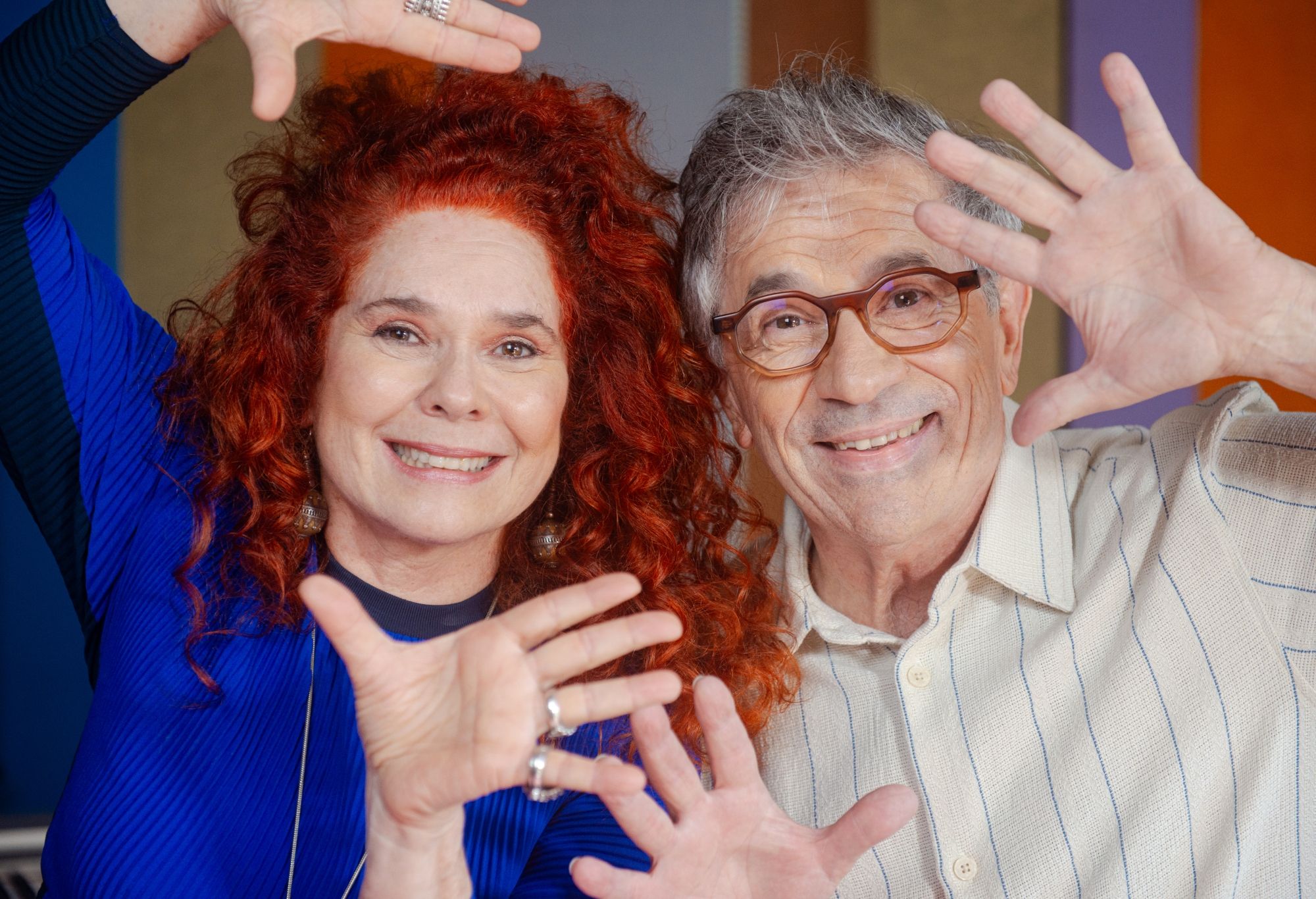 Sandra usa um vestido azul, tem cabelos cacheados e vermelhos e está com as mãos perto do rosto. Paulo, também com as mãos perto do rosto, aparece com uma camisa branca com listras azuis. Ele usa óculos de grau. Os dois são brancos e estão sorrindo. Sandra usa um vestido azul, tem cabelos cacheados e vermelhos e está com as mãos perto do rosto. Paulo, também com as mãos perto do rosto, aparece com uma camisa branca com listras azuis. Ele usa óculos de grau. Os dois são brancos e estão sorrindo.