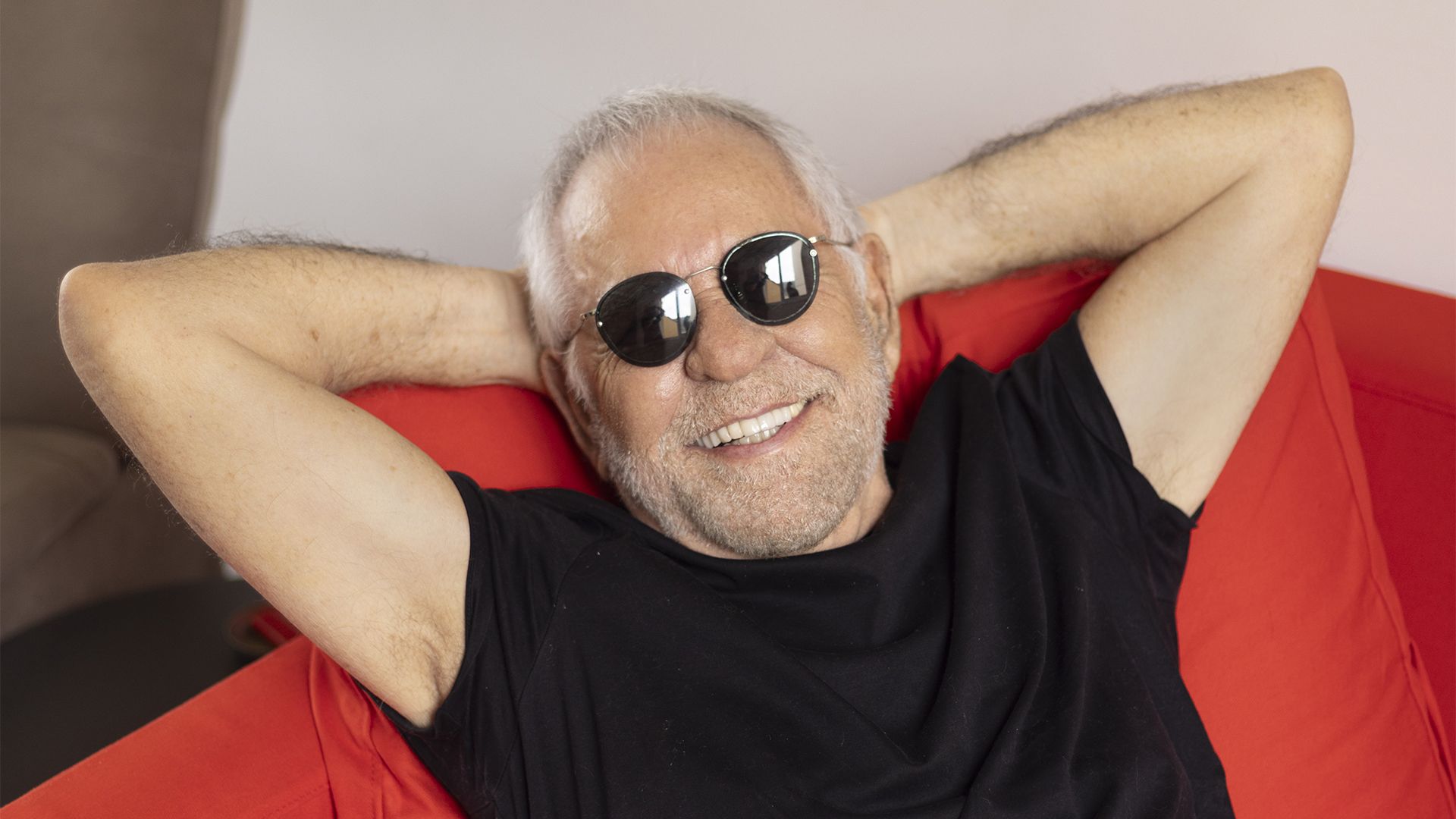 Fotografia colorida de Nelson Motta. Ele é um homem branco, calvo, de cabelos brancos e barba curta e branca. Nelson Motta veste uma camiseta preta, está sentado um um sofá vermelho, e tem os braços abertos com as mãos na nuca.