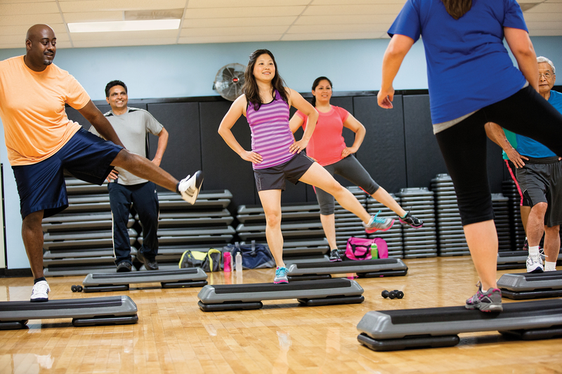 a group of adults perform a sideways kick move on a step bench in a group fitness class