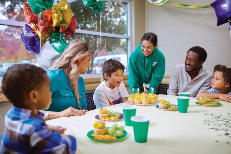 a group of adults and children sit around a table with cupcakes, balloons, and party decorations while a young boy blows out his candles on his cupcakes