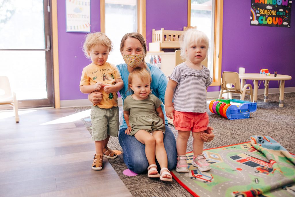3 preschool students smile with their teacher, Karissa, in the preschool classroom.