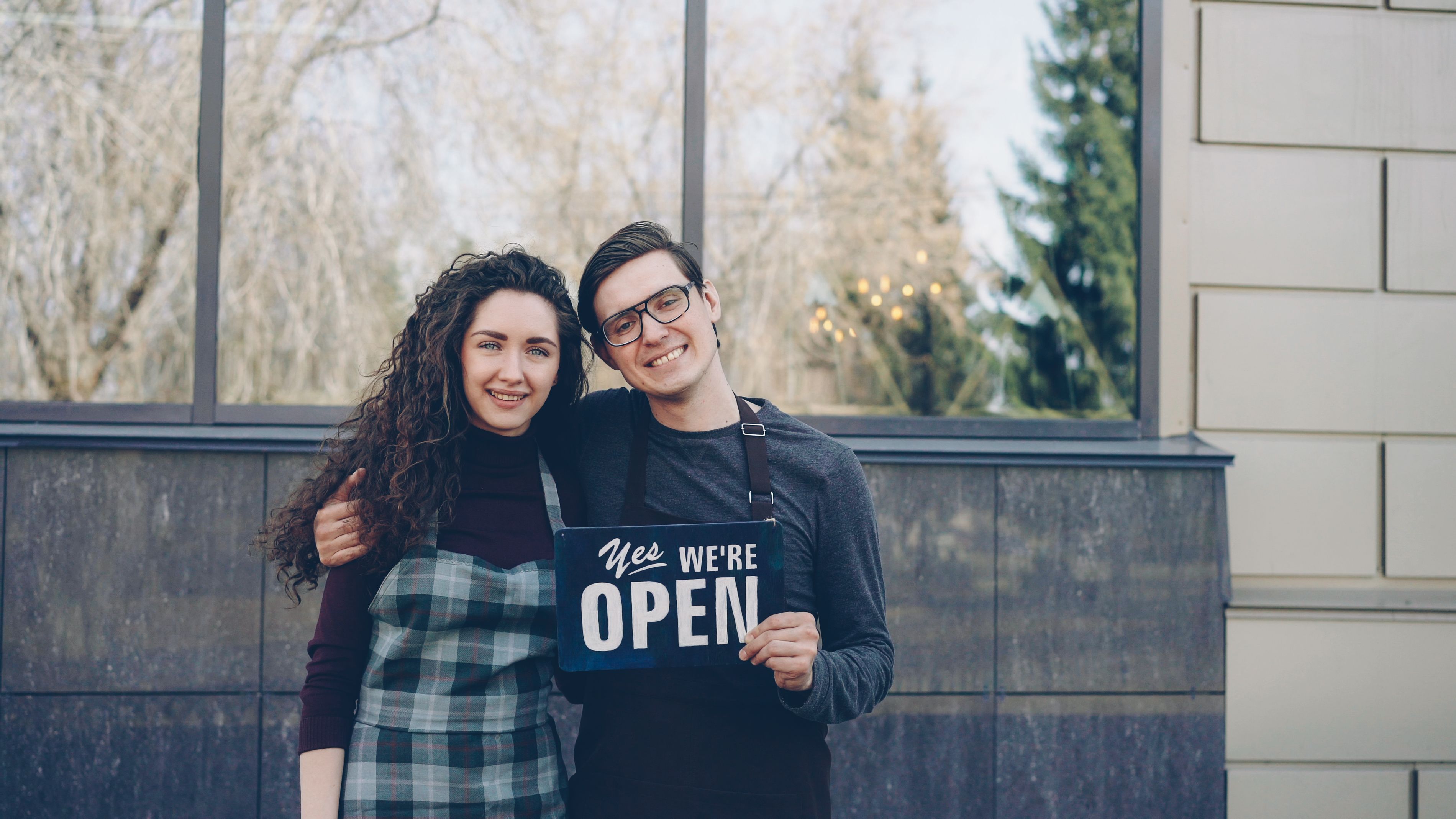 Two small business owners holding an open sign outside their storefront.