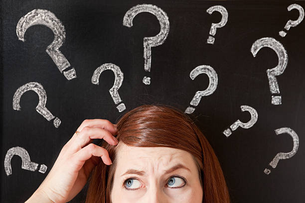 A confused scratching her head woman standing in front of a chalkboard with question marks written all over it.
