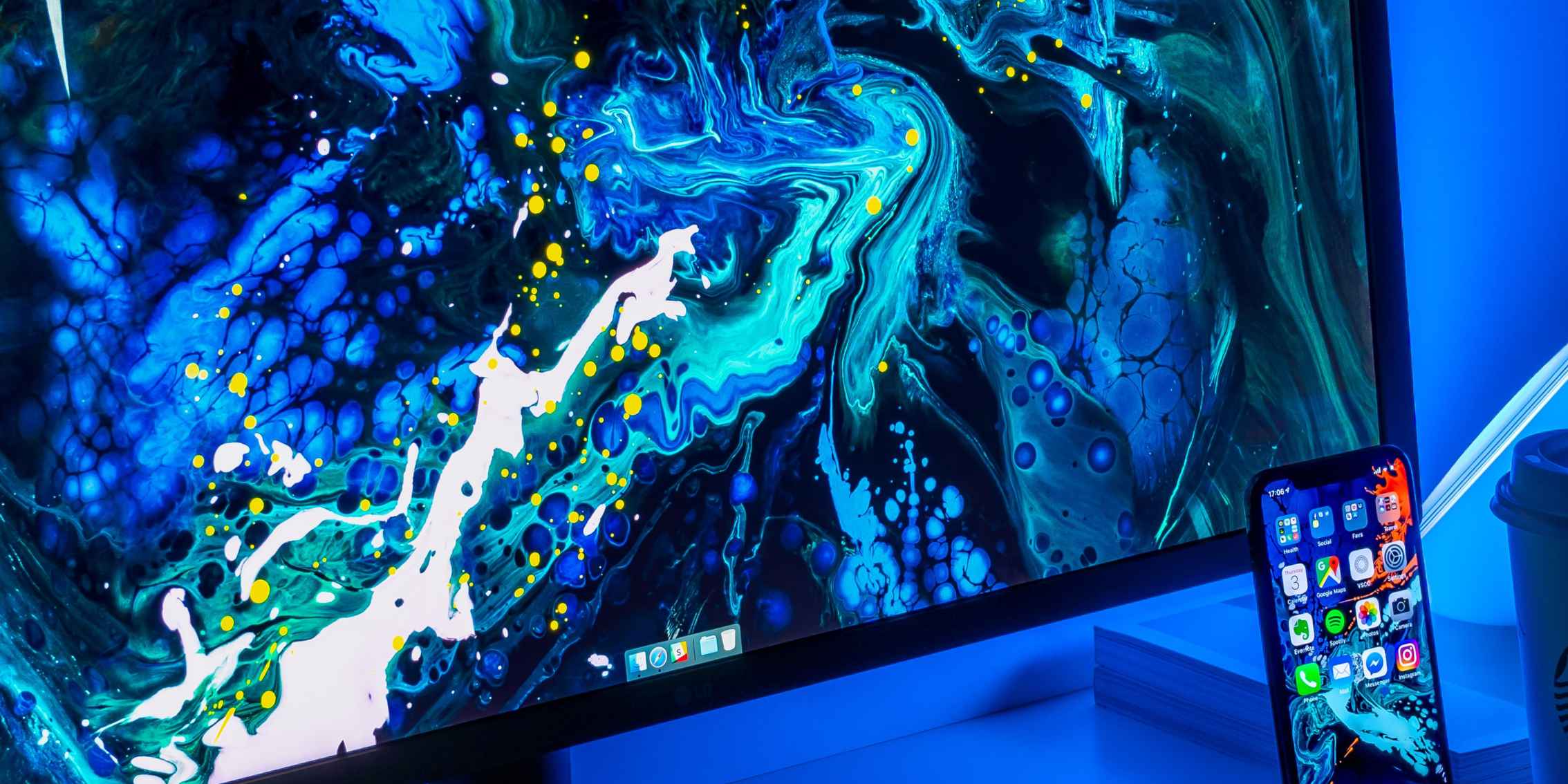 A computer monitor displaying a colorful abstract painting, with a smartphone and a cup of coffee on a nearby table. A computer monitor displaying a colourful abstract painting, with a smartphone and a cup of coffee on a nearby table.