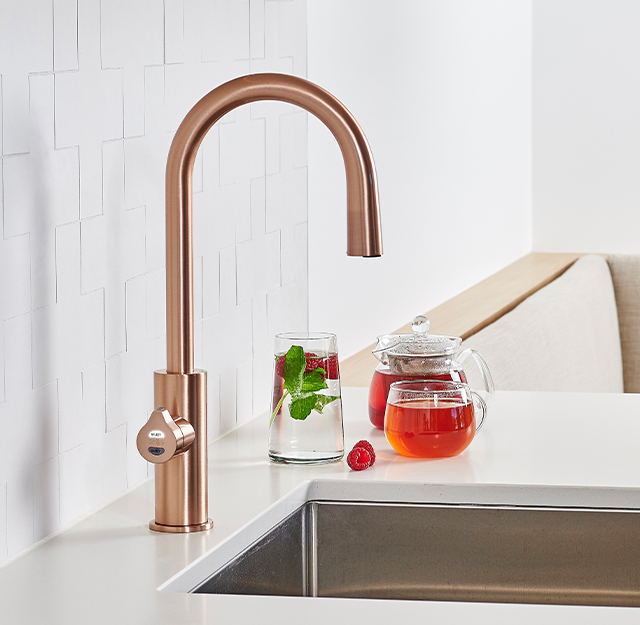 Rose gold kitchen faucet by Zipwater in an all white kitchen and silver sink. 1 glass of water with mint and raspberries, one clear tea pot and clear glass full of red tea.