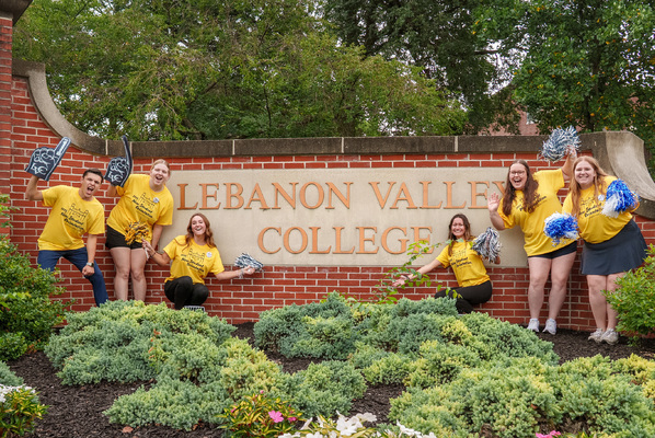 Four students cheer with pom-poms and foam fingers in front of a Lebanon Valley College sign