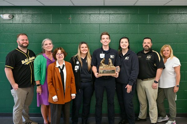 A group of people pose for a picture in front of a dark green wall while holding a trophy.