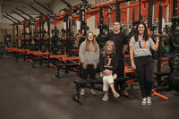A group of four people is seen in a weightlifting room in front of a long row of orange-and-black squat racks. 