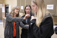 A woman holds a clear plastic measuring device on another woman's shoulder while a second woman watches. 
