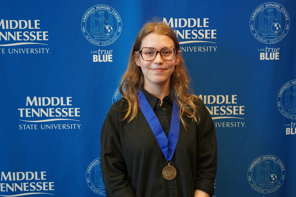 MTSU student with glasses smiling and wearing medal in front of backdrop