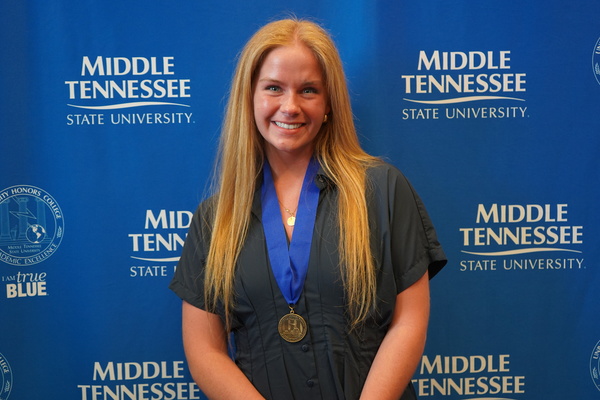 MTSU student smiles wearing thesis defense medal, standing in front of backdrop