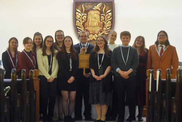 A group of nicely dressed students stands and poses in front of a large Elmira College Shield. 