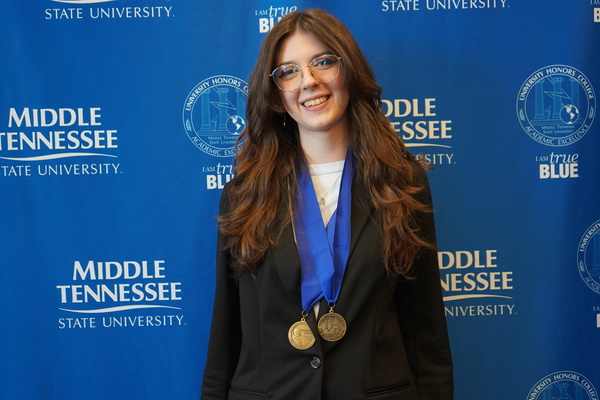 Smiling MTSU student just successfully defended Honors thesis, wearing medals in front of backdrop