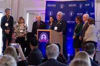 Eight people stand on stage while one delivers a speech at a podium