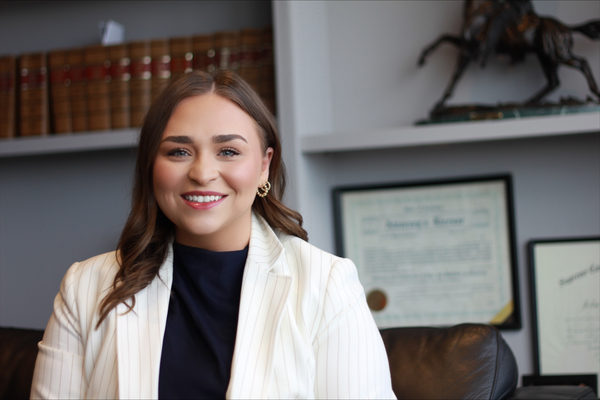 Kathryn Totty, in a black shirt and white blazer, is standing in front of a bookshelf in an office. 