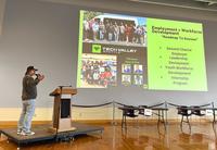 A person at a podium stands in front of a screen showing a presentation. The slide is about employment and workforce development.