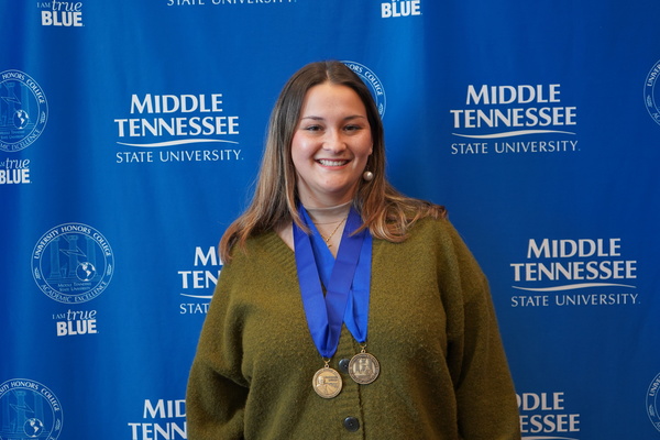 Smiling MTSU student just successfully defended Honors thesis, wearing medals in front of backdrop