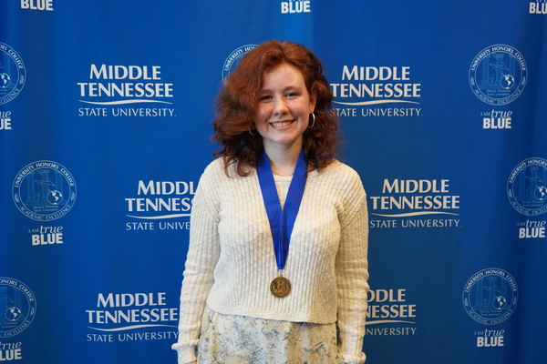 Smiling MTSU student just successfully defended Honors thesis, wearing medals in front of backdrop
