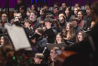 Crane School of Music students holding sheet music sing in performance, with the symphony orchestra's music stands in the foreground.