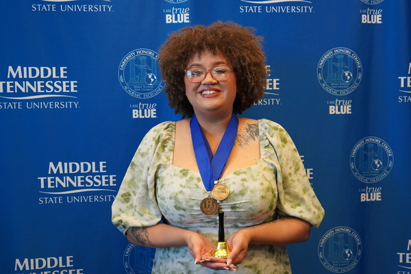 Smiling MTSU student just successfully defended Honors thesis, wearing medals in front of backdrop