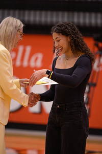 A woman in a yellow blazer hands a certificate and envelope to a woman in a dark dress in a gymnasium.