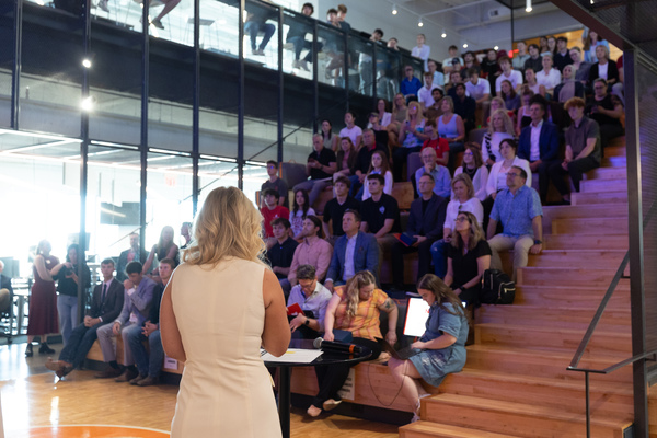 A woman speaks to a crowd seated on bleachers.