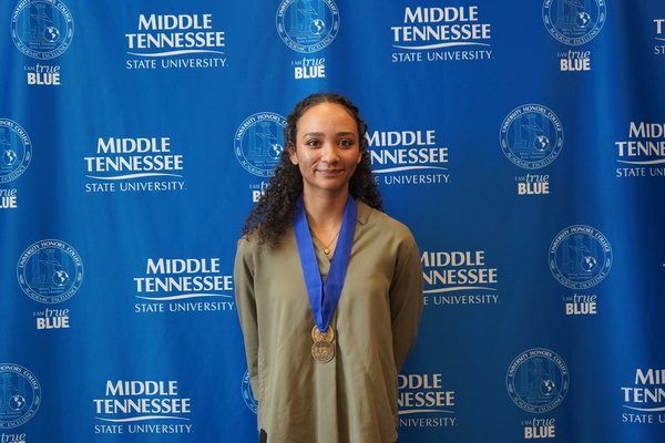 Smiling MTSU student just successfully defended Honors thesis, wearing medals in front of backdrop