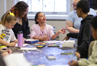 A girl gestures while working on a craft project, surrounded by college students in an art education program and classmates.