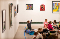 A child gestures at art on the wall of a gallery, while other children are seated and watching nearby.