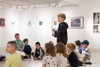 A boy stands and reads from a paper while visiting an art gallery. Fellow children are seated on the floor around him, looking at art and listening.