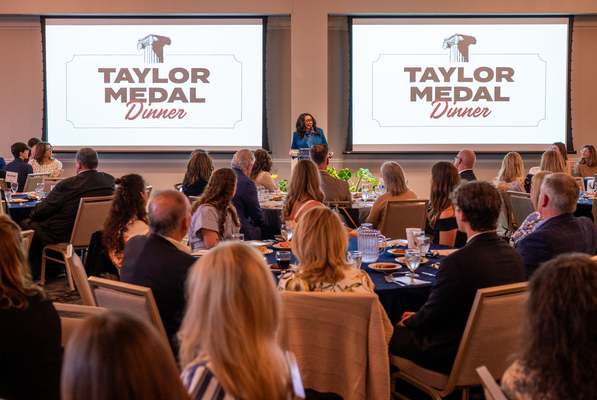 Guests of honor sit at tables and listen to a speaker who stands between screens with the words 'Taylor Medal Dinner.'