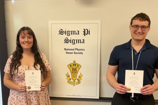 A pair of students hold certificates and stand in front of a white board with a sign for the Sigma Pi Sigma honor society.