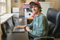 A student wearing a green shirt and a red ball cap works on a computer in the North Country Public Radio offices. 