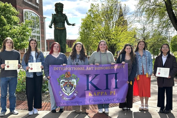 Eight students stand outside in front of an art sculpture and hold a banner that says Kappa Pi