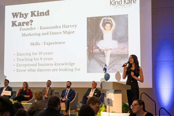 Woman presenting for panel of judges with slide on the way projecting Why Kind Kare?