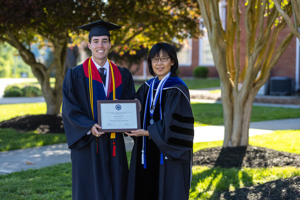 Student Jackson Estes and Professor Dr. Julie Tan pose in commencement regalia in front of two trees, with Estes holding his award certificate.