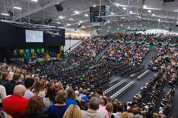 Gym with spectators in bleachers, graduates seating in chairs on floor and commencement stage.
