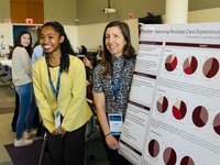 A student and professor in professional attire stand next to a research poster summarizing their work at a conference.