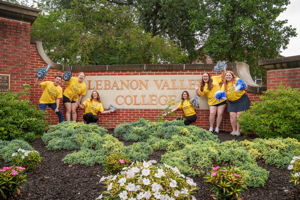 Students in yellow shirts cheer with pom poms near a brick wall sign for Lebanon Valley College