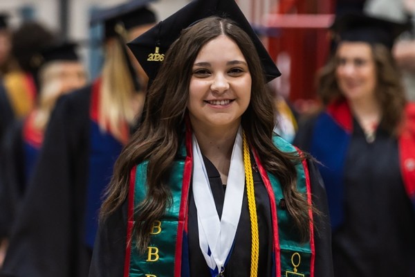 Smiling graduate in cap and gown with honor cords and medals walks during a commencement ceremony, with other graduates blurred in the background.